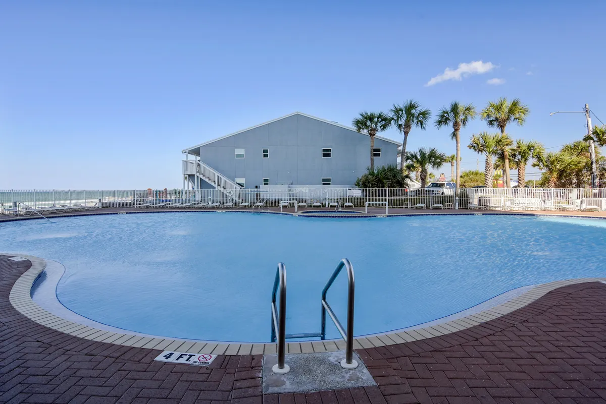 A sparkling swimming pool surrounded by lush palm trees and a building in the background, with areas perfect for sunbathing, swimming, and poolside social gatherings. The inviting ambiance highlights communal relaxation and opportunities for group fun.