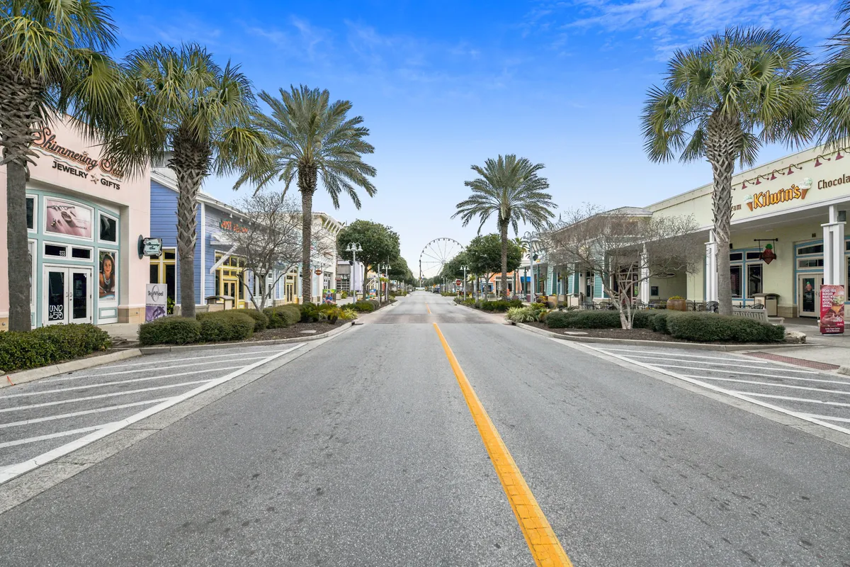 A lively outdoor scene featuring vibrant storefronts, towering palm trees, and a ferris wheel in the distance. The area is bustling with activity, ideal for shopping, dining, and strolling as a group, capturing the spirit of nearby Pier Park.