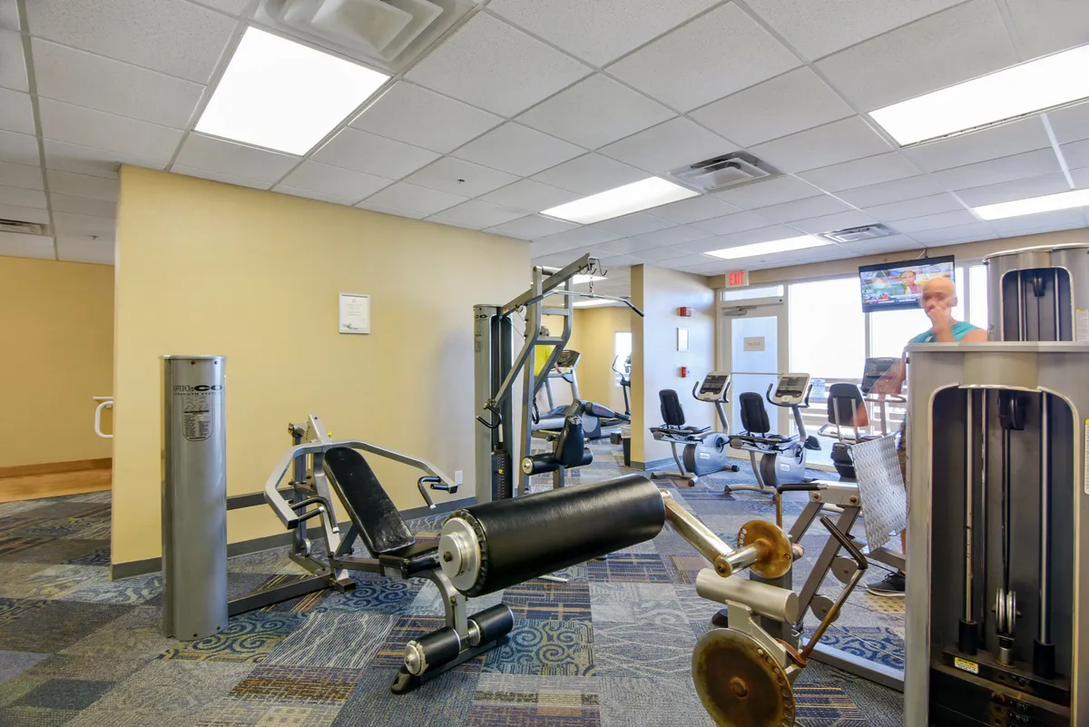 A modern fitness center filled with exercise equipment, large windows letting in natural light, and a television screen. The inviting open space suggests a motivational atmosphere for working out and socializing.