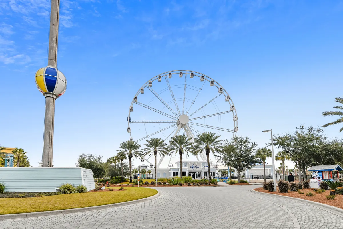 A bright, joyful exterior shot of Sky Wheel Panama City Beach, with the iconic Ferris wheel rising behind palm trees and the welcoming entrance in view. Families and children are seen strolling and preparing to ride, capturing the spirit of family-friendly adventure.