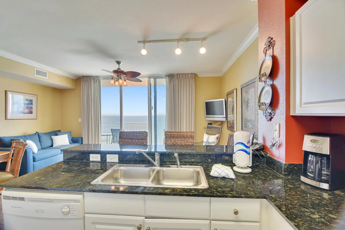 An open-concept interior that seamlessly blends a kitchen island, dining table, and living space. The kitchen overlooks the ocean through expansive windows, and a blue sofa anchors the living area. Decorative wall art and tasteful furnishings make the space feel inviting, perfect for hosting family meals or game nights with a view.
