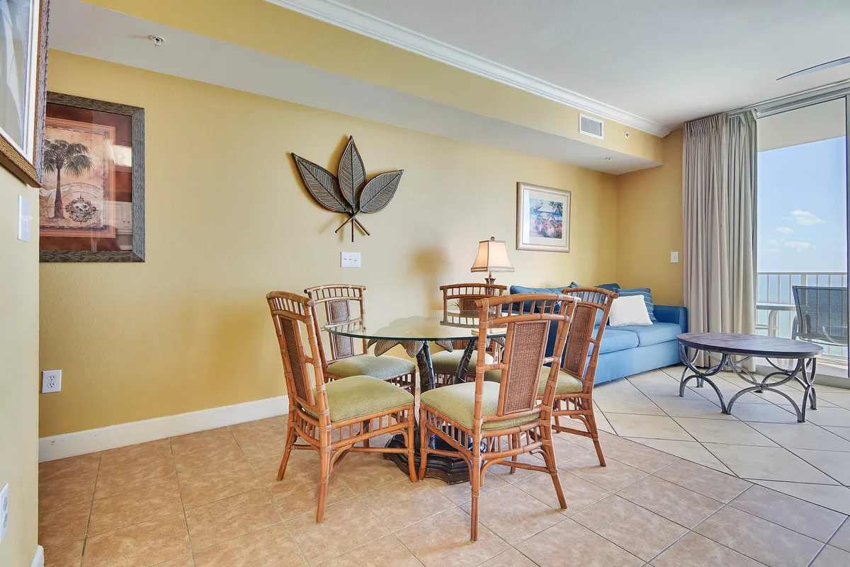 An inviting interior dining area featuring a glass table and rattan chairs, with a large window framing a beautiful ocean view. Morning light streams in, suggesting a perfect spot for breakfast while watching the sunrise over the water.