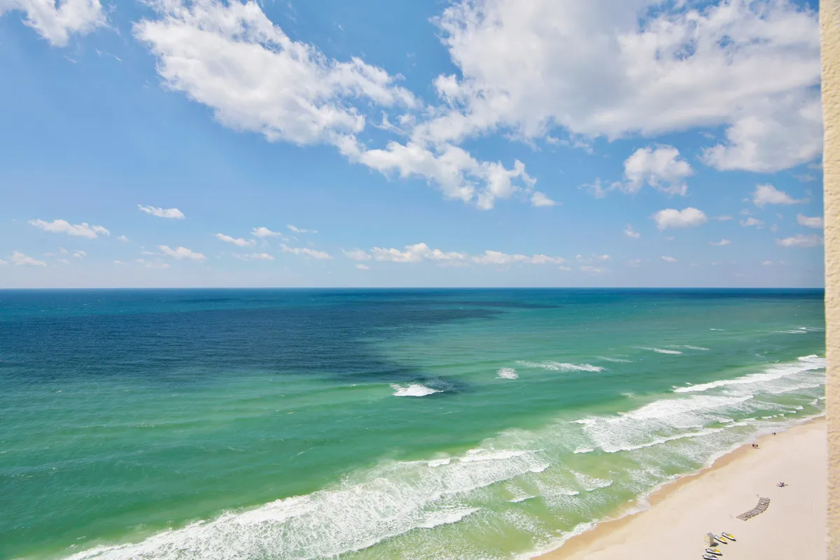 A sweeping view of the white sandy beach meeting the tranquil, blue-green Gulf waters beneath a sky dotted with fluffy clouds. The scene shows families enjoying the shoreline—swimming, sunbathing, and children building sandcastles. The vastness of the horizon emphasizes the resort’s prime beachfront location.