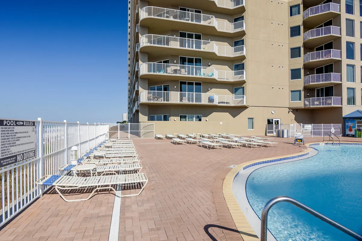 This image features a sparkling swimming pool with plenty of lounge chairs arranged around the edge and the resort's building facade in the background. The inviting blue water and sunny setting evoke relaxation and leisure.