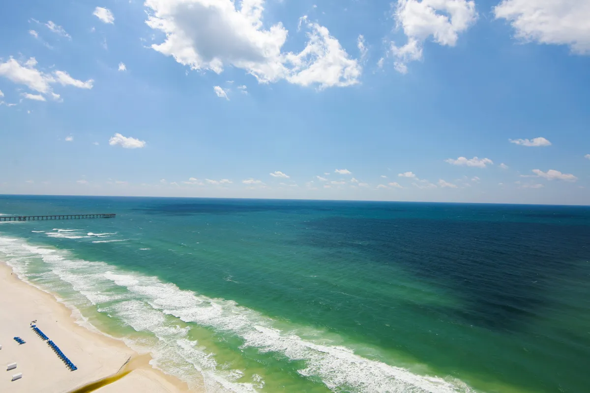 An expansive beach view showing the ocean, sandy shore, and a distant pier under a sky brushed with clouds. The restful scene invites beach walks, sunset watching, and waterfront relaxation.